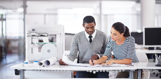 Two business owners are looking over design schematics while their 3D printer operates beside them.