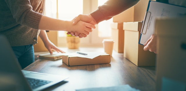 Two businesspeople shake hands at a desk to finalize a deal, surrounded by several packaging boxes and materials.