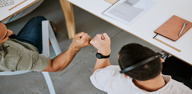 Two employees sitting side by side at a large L-shaped desk bump their fists together.