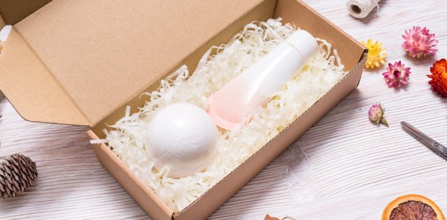 A brown cardboard e-commerce package filled with beige filler and two beauty products on a table covered in flowers.