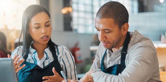 Two workers wearing aprons and sitting at a table with a laptop on it go over details in a notebook while talking.