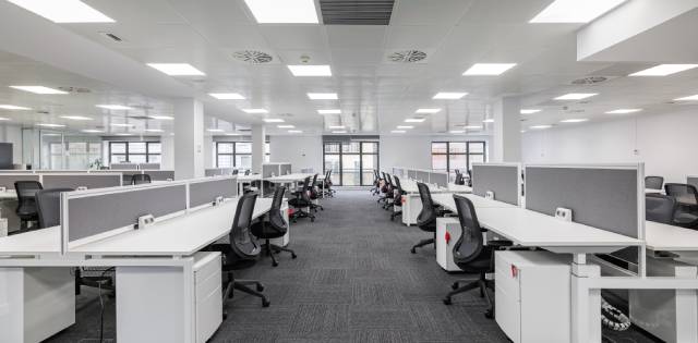 A modern office that has adjustable desks and wide pathways. The desks are white and the chairs are black.