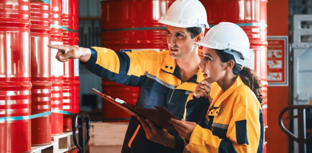 A man and woman in hard hats and safety gear pointing at and discussing a room full of shiny red barrels.