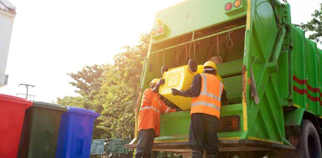 Two men neatly dressed in orange safety vests and hardhats empty a bright yellow bin into a garbage truck.