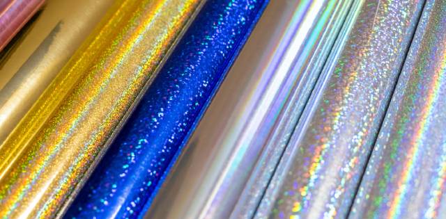 Various rolls of holographic and glittery vinyl media of different colors, including gold, blue, and silver.