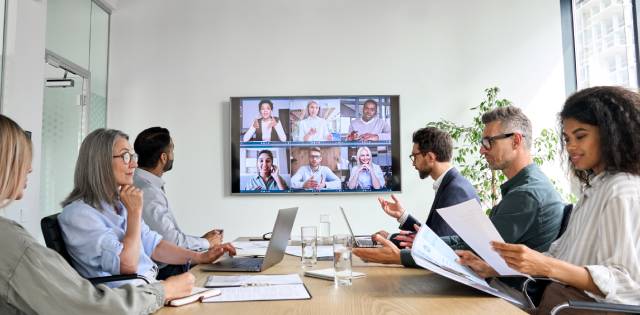 People sitting at a conference table look at their laptops and papers. A TV screen shows additional participants.