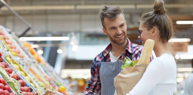 A friendly produce manager assists a female shopper who's holding a paper grocery bag in the produce section.