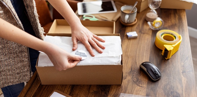 A woman's hands, up close, as she places a sticker on a folded piece of packing paper in a cardboard box.