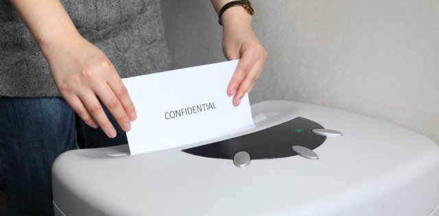 A person standing behind a paper shredder, feeding in a white document that reads "confidential" on the front.