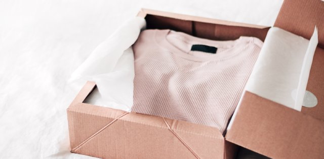 A tan-colored waffle-textured sweater is neatly folded in a brown cardboard box. There is white tissue paper inside, too.