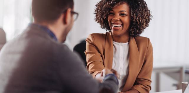A woman in a brown blazer smiling widely as she shakes the hand of a man across the table with his back to the camera.