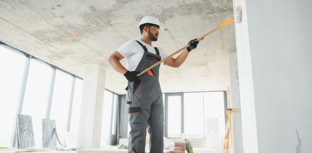 A man wearing gray overalls and a white hard hat is standing in a business mid-renovation. He's painting the wall white.