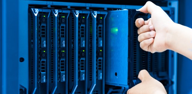 A person’s hands pulling a hard drive out of a server cabinet with small LED lights on it in a data center.