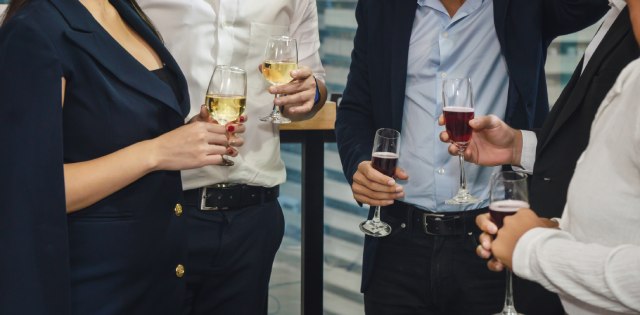 Five people in business attire hold wine glasses and mingle. They stand together in a small circle.