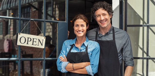 A man and woman wearing aprons and smiling stand in the doorway of a business with an open sign facing outwards.