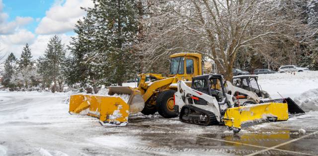 Three compact tractors with snow plows parked in a snowy parking lot, waiting to begin the snow removal process.