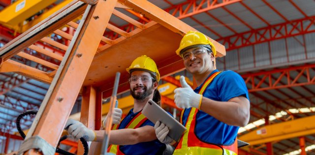 Two men wearing personal protection equipment give a thumbs-up as they work in a warehouse. They're both smiling.