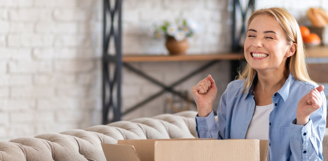 A woman excitedly gesturing with a big smile after opening a cardboard package. She is sitting in her living room.