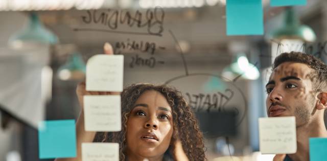 Two business partners brainstorm branding ideas while pointing at a glass board covered in colorful sticky notes.