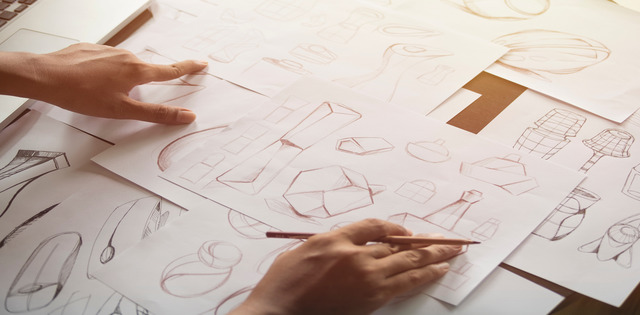 Close-up of a person's hands sketching the concepts of various products on sheets of paper with a pencil.