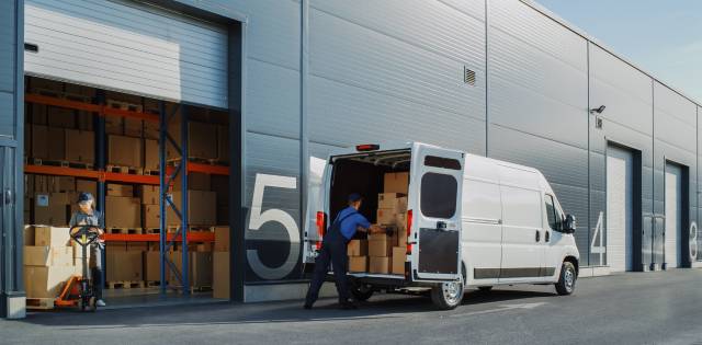 Two workers outside a large warehouse use a pallet to load up a small truck with cardboard boxes on a sunny day.