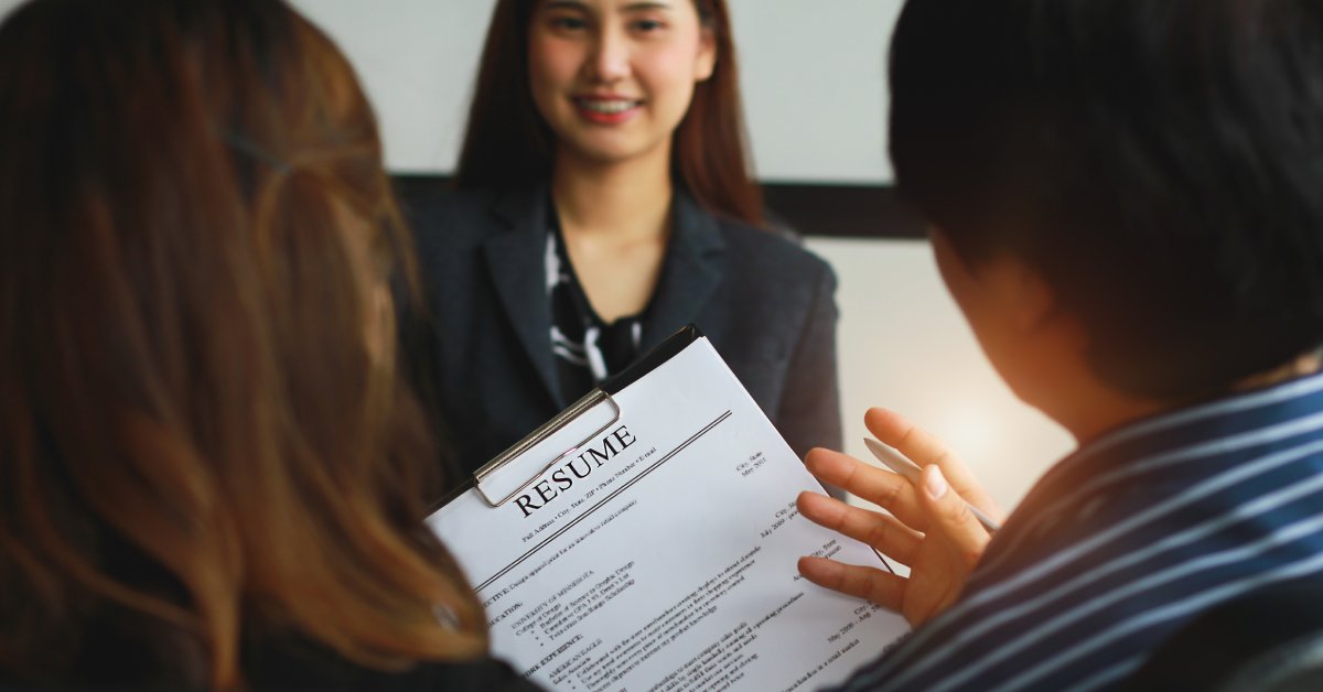 A woman is seen smiling between two people who are interviewing her. One holds her resume and gestures.