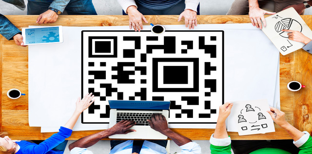A bird's eye view of a table with a large QR code printed on white paper in the center. Six people sit at the table.
