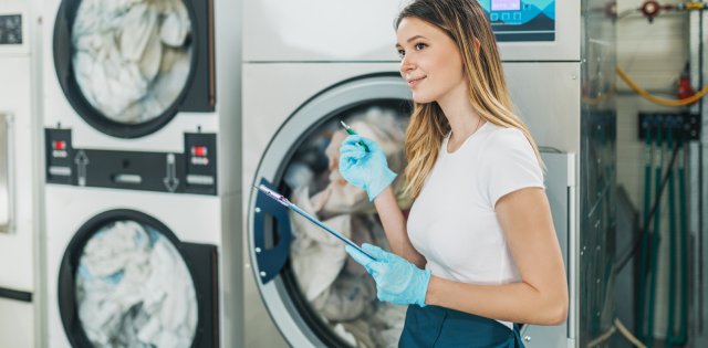 A woman wearing blue gloves and an apron leans against an industrial washing machine while holding a clipboard.
