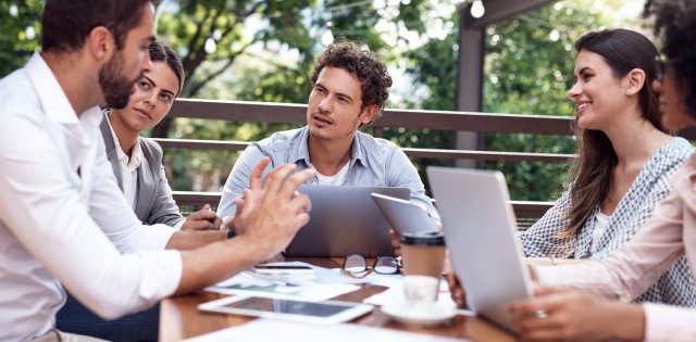 Business professionals holding a meeting outside. There are laptops, tablets, and paperwork on a wooden table.