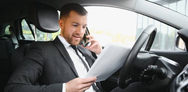 A business professional sitting in the driver seat of the car. He is talking on his cell phone and holding papers.