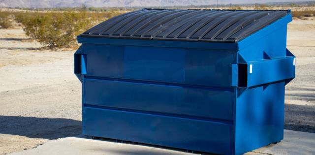 A rectangular blue dumpster is sitting on a flat concrete surface. The black lid on top of it is closed to cover the trash.