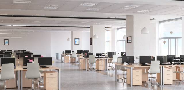 A row of desks, chairs, and office drawers. There are monitors on top of the desks and large windows in the office.