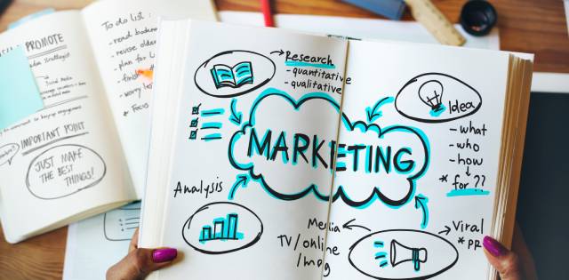Closeup of a woman’s hands holding an open notebook that says “marketing” in the middle with various notes surrounding it.
