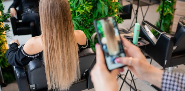 The blurred hands of a stylist using a phone to take a photo of the hair of a female client sitting facing a mirror.