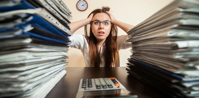 A woman sits at a desk with her hands on her head. Two large stacks of documents and a calculator are in front of her.