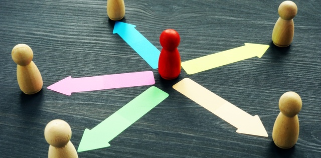 A small red wooden figure in the center of five other plain wooden figures. Colorful arrows point inward at the red figure.