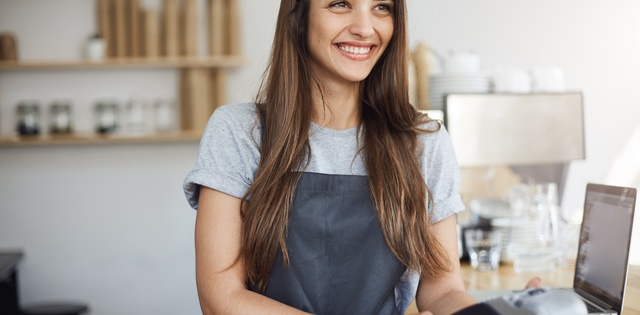 A young woman wearing a gray apron stands behind a counter holding a card reader to a customer out of frame.
