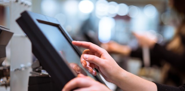 Hands interacting with a touchscreen point-of-sale system, with another person and blurred surroundings in the background.