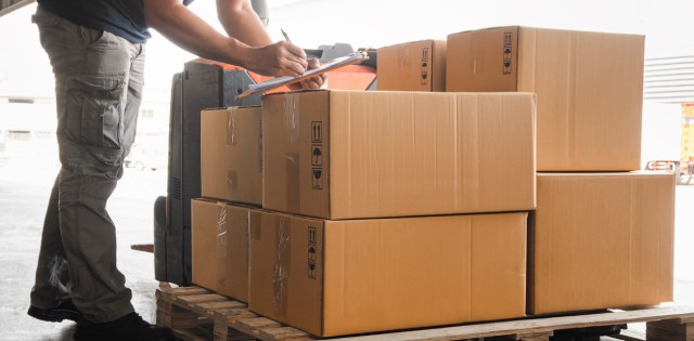 A man leans over a pallet of cardboard boxes containing inventory while signing a form on a clipboard.