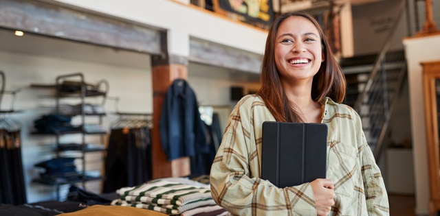 A woman in a brown plaid shirt smiles while holding a tablet, surrounded by neatly arranged clothes in a store.