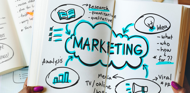 Close-up of a woman’s hands holding an open notebook with “marketing” written in the middle and brainstorming ideas.