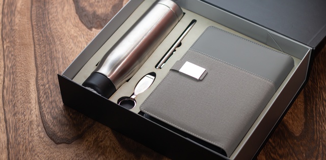 A corporate gift package with a stainless steel water bottle, pen, key chain, and journal in a box on a wooden table.